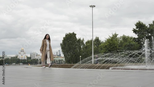 A girl with long hair and a brown trench walks along the fountain on a summer warm evening. High quality 4k footage