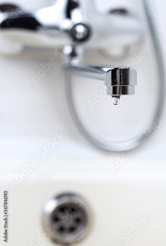 Faucet with a drop of water against a white sink. Water leaking and saving. Selective focus.