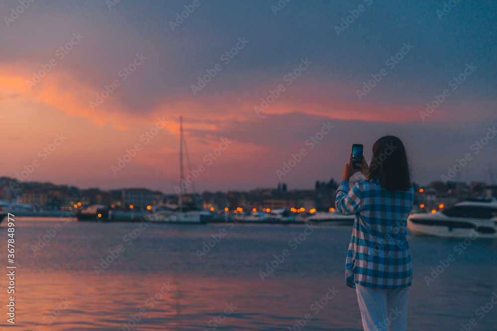 Fototapeta premium woman taking picture of the sunset over sea dock on her phone