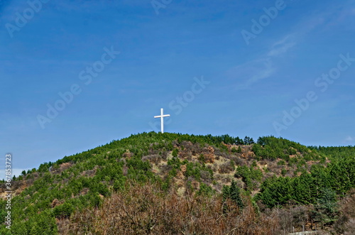 A majestic wooden cross rises over a hill overgrown with coniferous and deciduous trees against the blue sky near Blagoevgrad, Bulgaria  