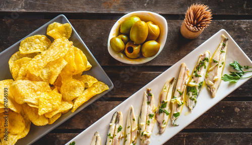 Three dishes with potato chips, picked anchovies and green olives on a dark wooden table with toothpicks. Typical Spanish snacks to eat in a bar. 