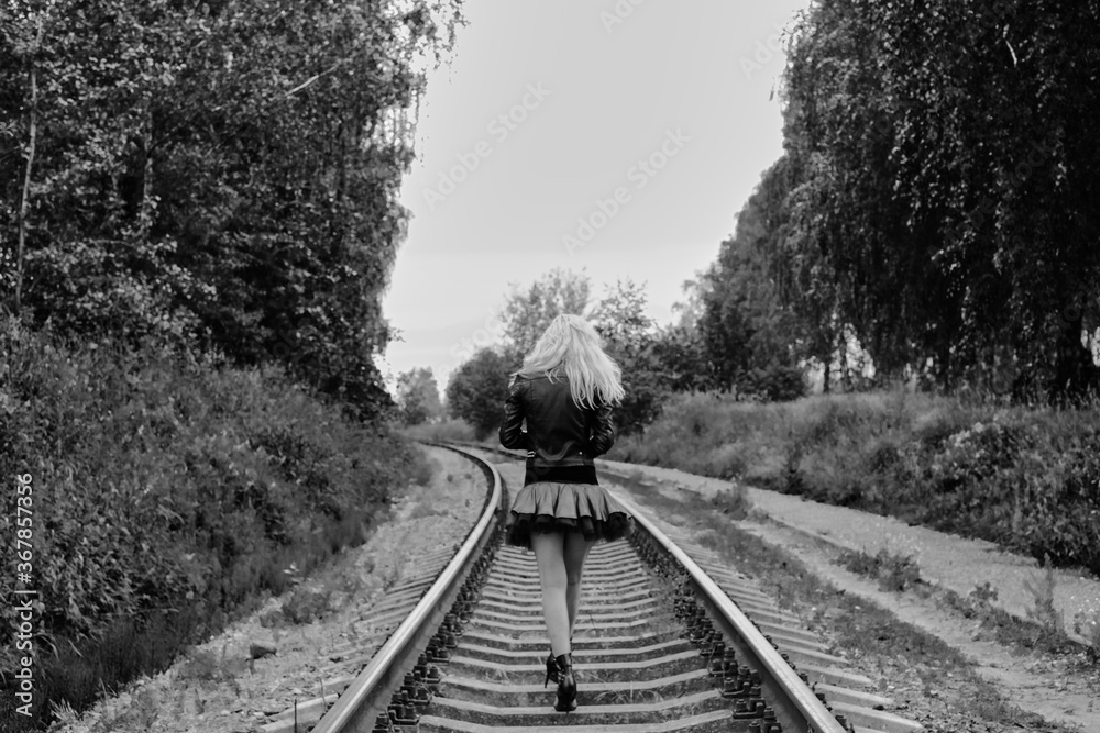 Blonde girl in a skirt and a leather jacket walking along the railway tracks black and white photo