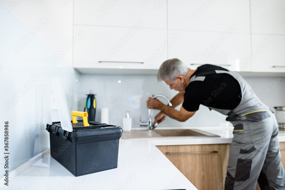 For all things. Aged repairman in uniform working, fixing broken kitchen tap using adjustable wrench. Repair service concept For all things. Aged repairman in uniform working, fixing broken kitchen tap using adjustable wrench. Repair service concept