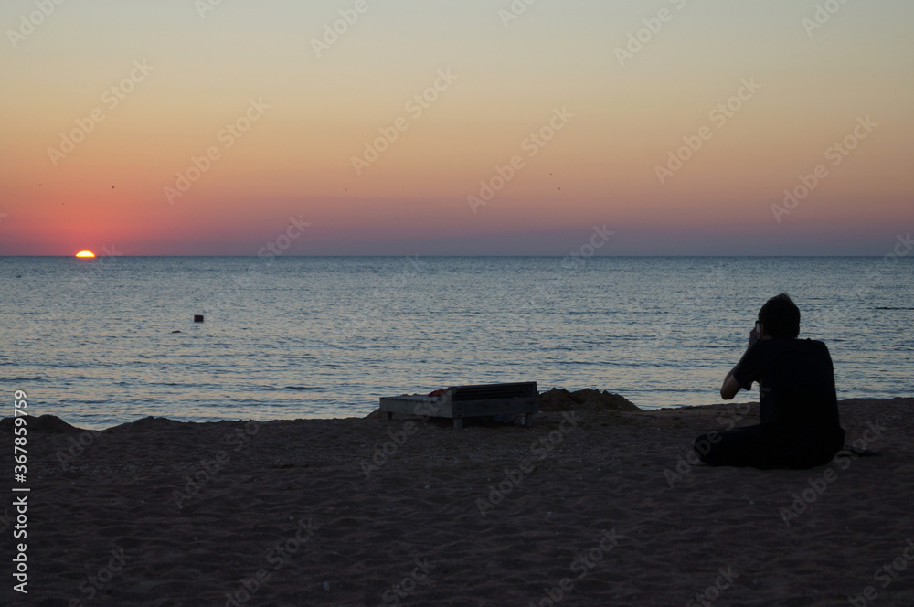 Silhouette of a man sitting on a deserted beach and photographing the sea during sunset