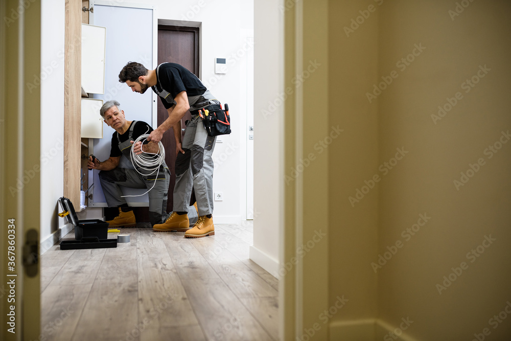 Work for possibilities. Full length shot of aged electrician, repairman in uniform working, installing ethernet cable or router in fuse box while his young colleague bringing cable