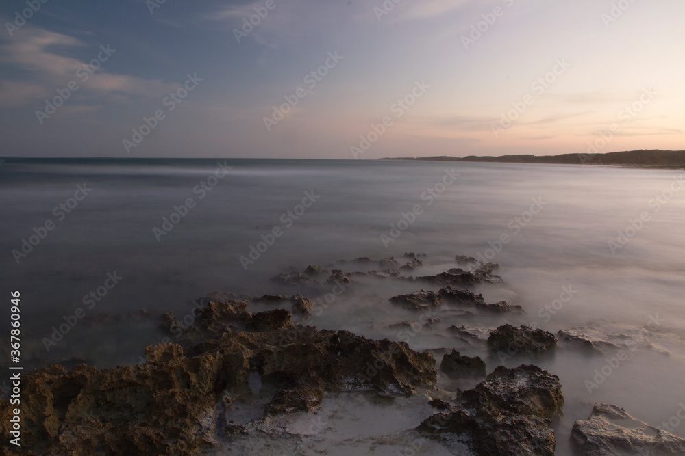 Long exposure. Serene tropical landscape at nightfall. The beach, rocks, ocean, shore, and sea waves with a smoky blurred effects with beautiful sunset colors. 