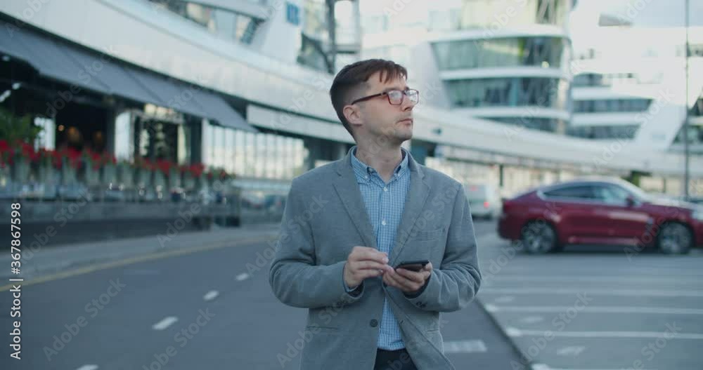 Young male businessman walks modern street and writes texting colleagues on mobile. An important negotiation. Against the background of a modern street with glass buildings and roads Young male businessman walks modern street and writes texting colleagues on mobile. An important negotiation. Against the background of a modern street with glass buildings and roads