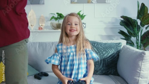 Little sweet daughter playing video game on TV console while her father swinging son upside down making obstacles. Living room fun. Home entertainment.