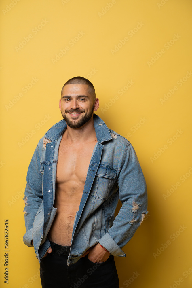 Fototapeta premium a smiling man with a naked body with a black beard and a fashionable barbershop haircut is photographed in a photo Studio, with a blue denim jacket thrown over the top