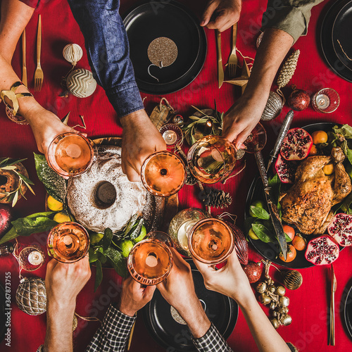 Friends celebrating Christmas. Flat-lay of people clinking glasses with rose wine over festive table with red cloth with roasted chicken, bundt cake, fruit, decorations, top view, square crop