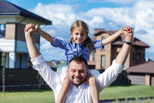 Photo of a dad holding his little daughter with love on his shoulders against the background of his new house. Happy family, child life insurance, baby care. Investment in children. Happy home photo