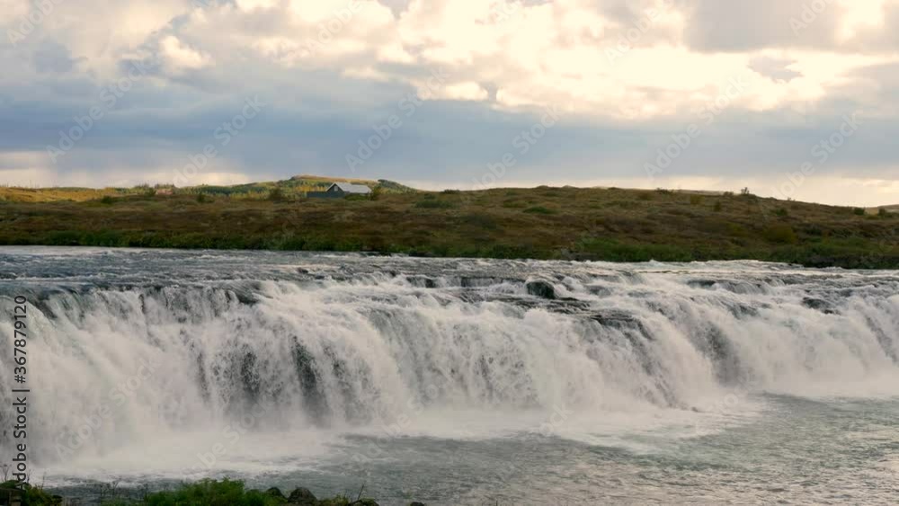 A raging waterfall on a river in Iceland. A house on a sunny hill in the background. A raging waterfall on a river in Iceland. A house on a sunny hill in the background.