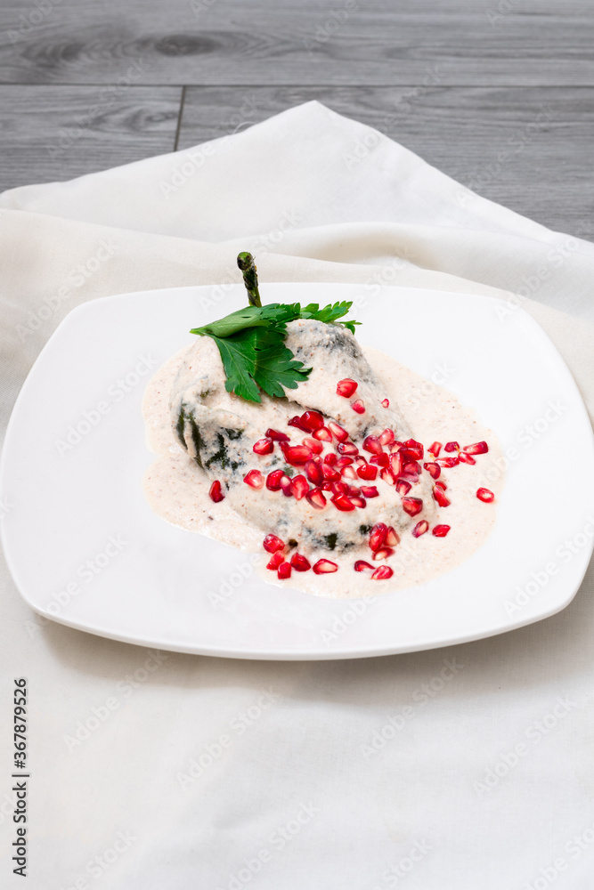 Chile en Nogada, Mexican dish that dates back to the colony, poblano pepper with a cream of walnut, with grains of pomegranate, in a white plate that is on a gray wooden table, next to it is a napkin