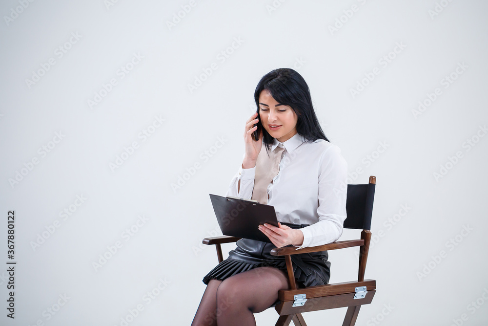 Successful business woman in a white shirt sitting on a chair with documents and talking about the business on the phone. Successful business woman in a white shirt sitting on a chair with documents and talking about the business on the phone.