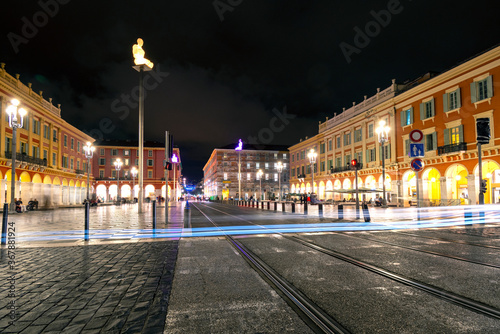 Fototapeta Naklejka Na Ścianę i Meble -  Car headlights make light trail from long exposure as the cross tram tracks late at night in Place Massena in the historic city of Nice, France.