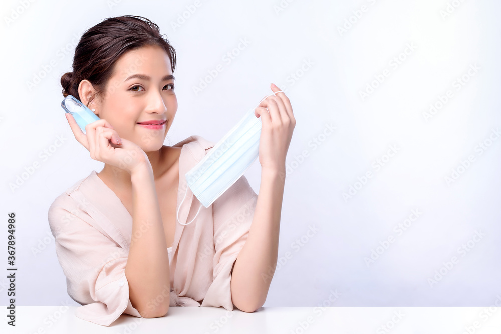 Portrait of beautiful charming young Asian woman posing with surgical mask or face mask to protect from corona virus and sitting with white table on white background, health care protection Covid-19 Portrait of beautiful charming young Asian woman posing with surgical mask or face mask to protect from corona virus and sitting with white table on white background, health care protection Covid-19