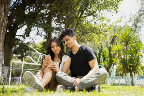 Hispanic young couple sitting in the park using smart phone on a sunny day - young people making video call with their phone © Fernanda