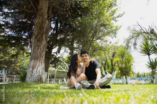 Young Hispanic couple in love sitting on the grass hugging and having fun - couple strolling in a natural park on a sunny day