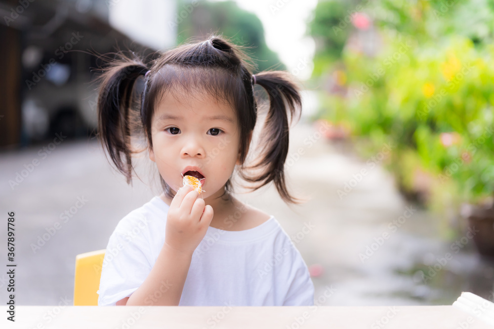 Cute little girl bunches hair and eating oranges into her mouth. Child wore a white shirt sitting in front of the house. Background is a small public road. Asian children eat fruit, aged 3 years old.