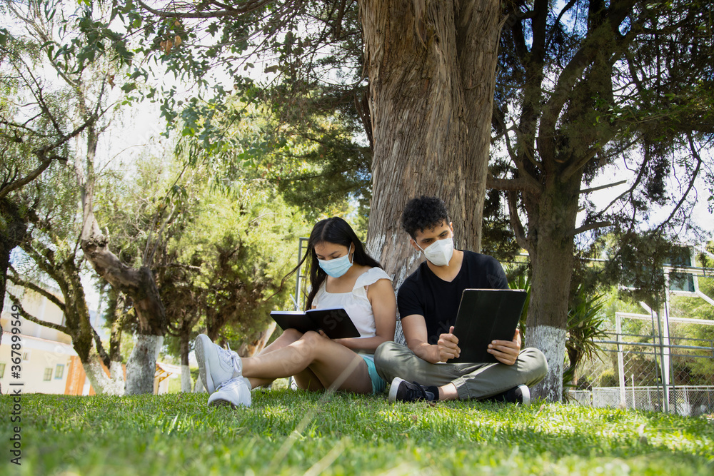 Young Hispanic couple sitting reading in the park with mask - young people studying with an electronic tablet and a book in a natural park - social distancing - general shot