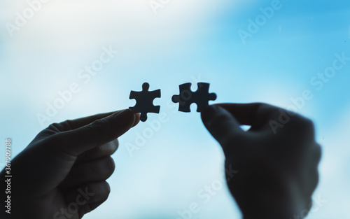 Closeup image of hands holding and putting a piece of white jigsaw puzzle together with blue sky background