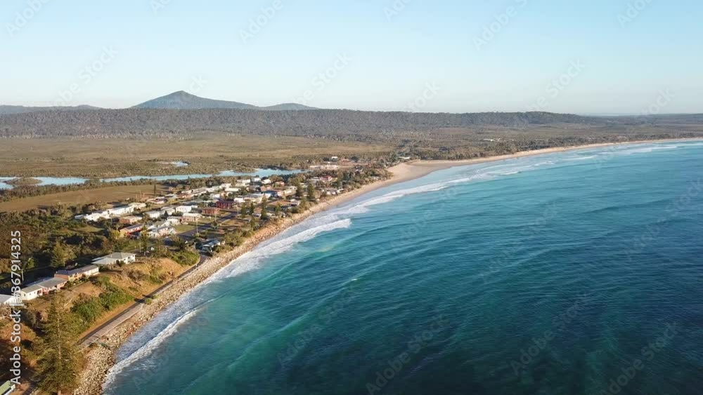 Aerial Drone On Sunny Day Over Blue Ocean And Coastal Seaside Town With Trees And Mountain In The Distance Aerial Drone On Sunny Day Over Blue Ocean And Coastal Seaside Town With Trees And Mountain In The Distance