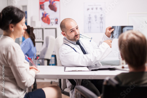 Doctor pointing at knee injury on x-ray to disabled senior woman in wheelchair and daughter during consultation in hospital office. Patients listening medic while explaining diagnosis.