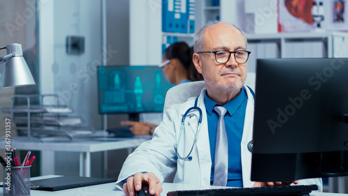 Elderly doctor working on PC in modern private clinic with glass walls. Hospital professionals in health industry staff at work in medicine medical practitioner specialist in unifor at desk job