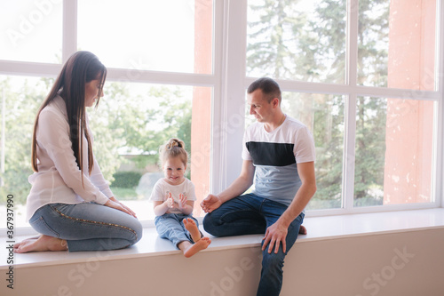 Little girl with mom and dad sits by the big window in studio. Family photosession