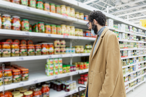 Young man in the supermarket. Shelves with a large selection of canned food. Brunette with a beard in a medical mask in a beige coat.