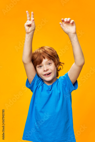 Cheerful boy hands raised up blue t-shirt yellow background red hair