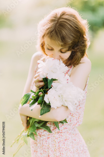 Beautiful young Caucasian blond woman in light dress holding bouquet of white peonies  walking in summer field or garden in sunset. Woman with flowers outdoors