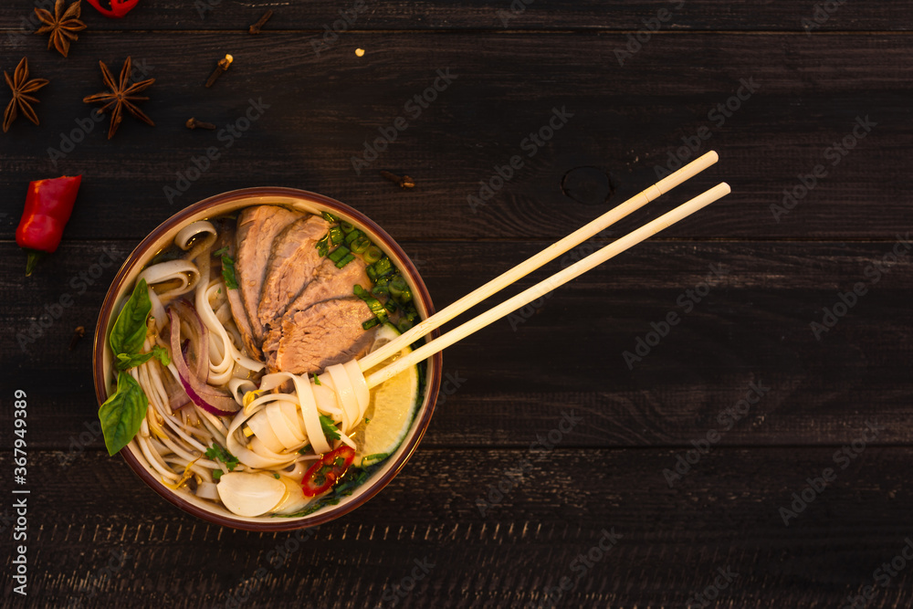 Vietnamese traditional spicy pho bo soup with rice noodles, beef slices, chili pepper, cilantro, onion, star anise on a brown wooden background. Chinese chopsticks in a plate. Flat lay. Vietnamese traditional spicy pho bo soup with rice noodles, beef slices, chili pepper, cilantro, onion, star anise on a brown wooden background. Chinese chopsticks in a plate. Flat lay.