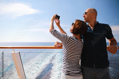 bald man and brunette woman traveling on cruise liner ship and taking pictures from smartphone on balcony 