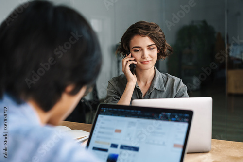 Image of pleased woman talking on cellphone while working with laptop