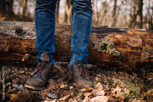 close up photo of human legs in old leather boots and blue jeans standing on the ground with dried leaves