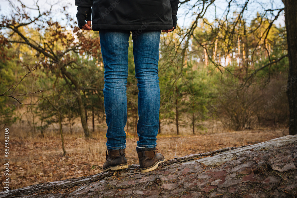 Young hiker in blue jeans and brown leather boots, standing on the tree trunk Young hiker in blue jeans and brown leather boots, standing on the tree trunk