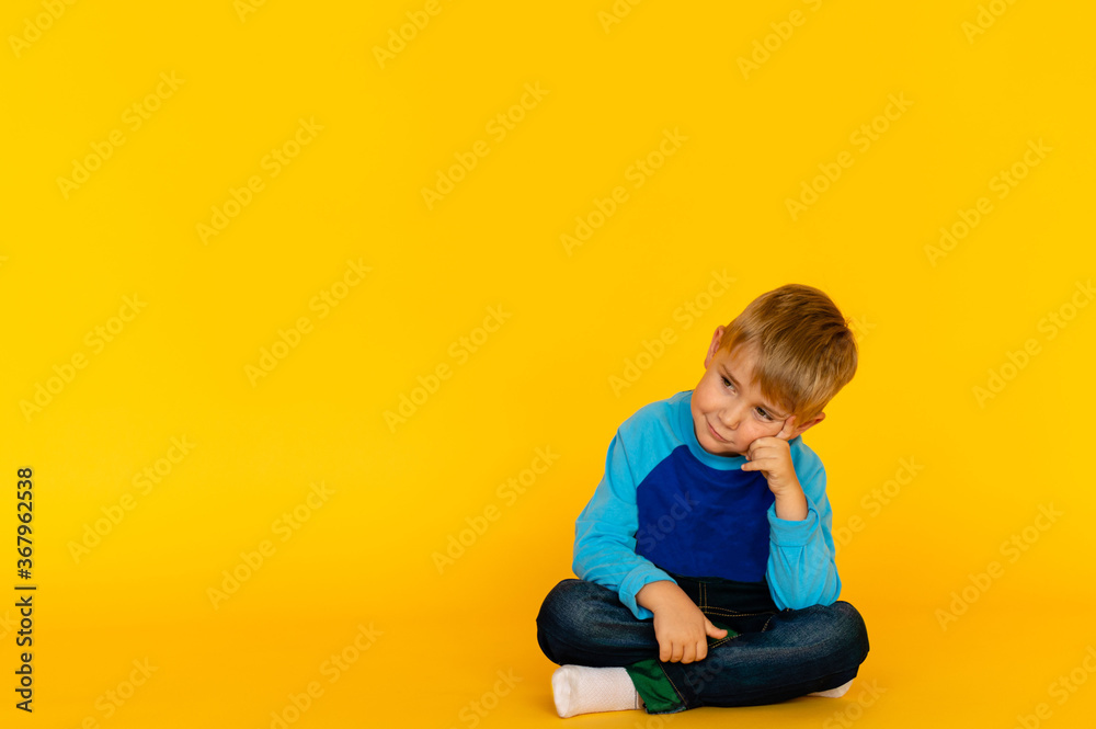 The boy is blond in bright blue clothes on a yellow background. The thoughts of the child. Thinker. Childhood. Beautiful background with the ability to insert text. The boy is blond in bright blue clothes on a yellow background. The thoughts of the child. Thinker. Childhood. Beautiful background with the ability to insert text.
