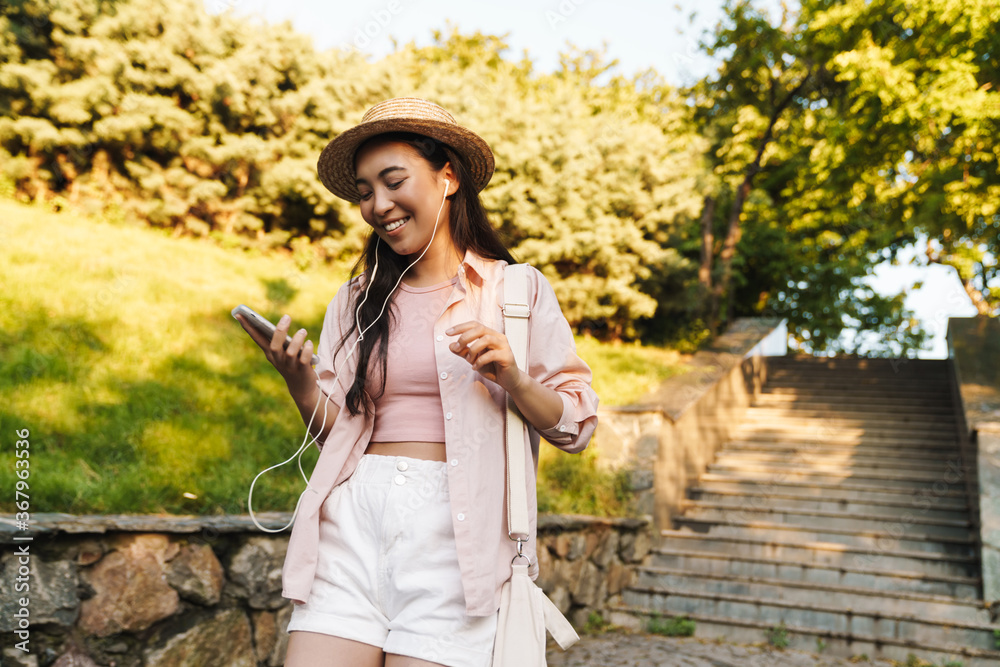 Obraz premium Image of cheerful asian girl in hat using mobile phone and earphones