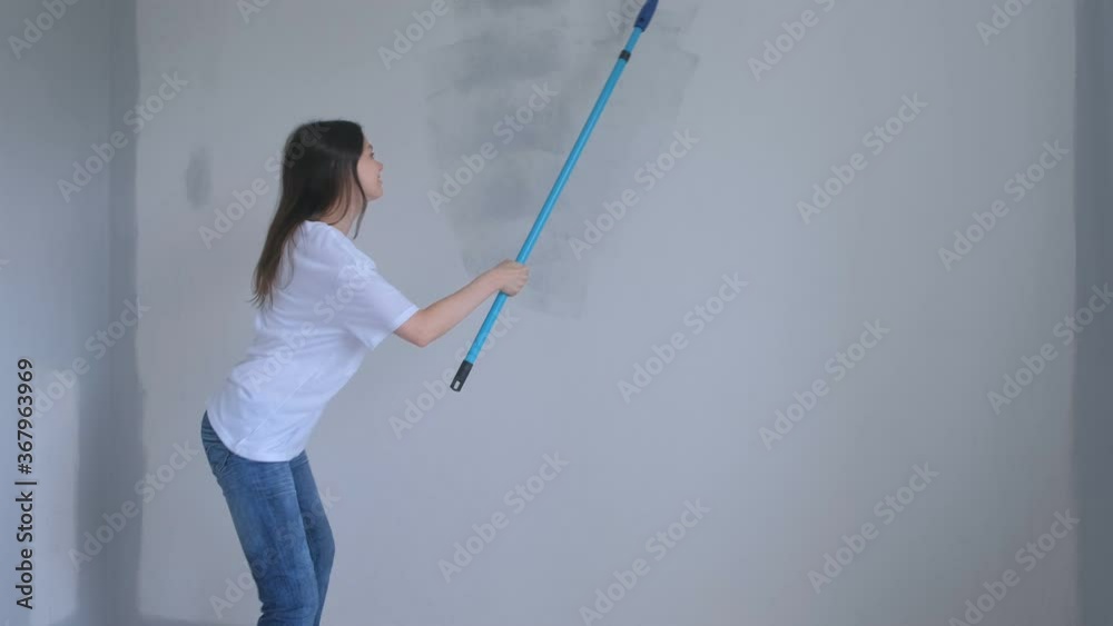 Young girl painter is painting wall in gray using paint rollers and dancing. Independent woman making renovation in new apartment, flat, room. Construction and finishing work in house herself. Young girl painter is painting wall in gray using paint rollers and dancing. Independent woman making renovation in new apartment, flat, room. Construction and finishing work in house herself.