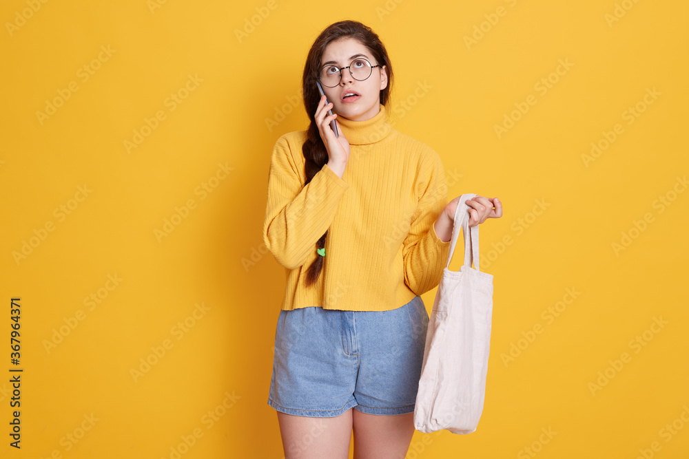 Young woman with cotton shopping bag talking on mobile phone while looking up, lady holding eco bag in hands, wearing yellow sweater and jeans short. Young woman with cotton shopping bag talking on mobile phone while looking up, lady holding eco bag in hands, wearing yellow sweater and jeans short.