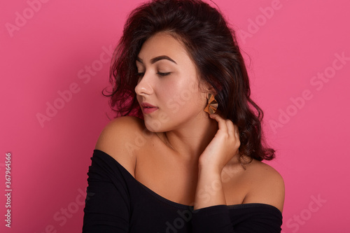 Portrait of brunette woman with wavy hair posing with hands on neck and looking aside, wearing black shirt with bare shoulders, standing isolated over pink background.