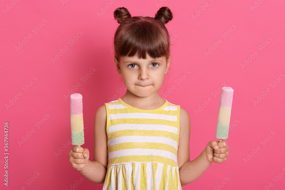 Portrait of little girl holding two fruit ice creams, posing with serious facial expression against pink background, dresses white and yellow attire. Portrait of little girl holding two fruit ice creams, posing with serious facial expression against pink background, dresses white and yellow attire.