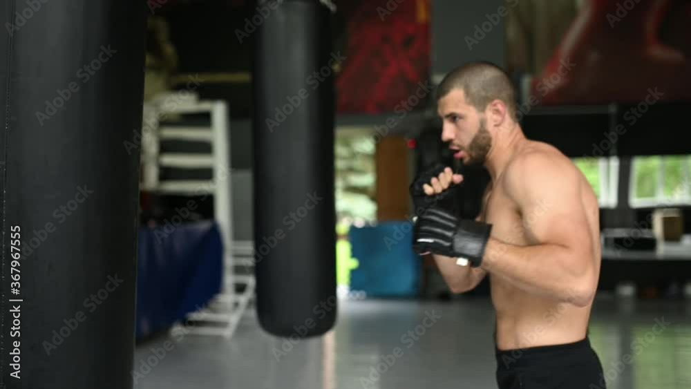 Fighter Practicing Some Kicks With Punching Bag. Kick, punching bag on dark background. Black punching bag weighs at the gym Fighter Practicing Some Kicks With Punching Bag. Kick, punching bag on dark background. Black punching bag weighs at the gym