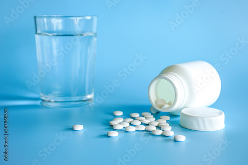 Close up of teblets pills spilling from pill bottle with a glass of water on blue background  photo