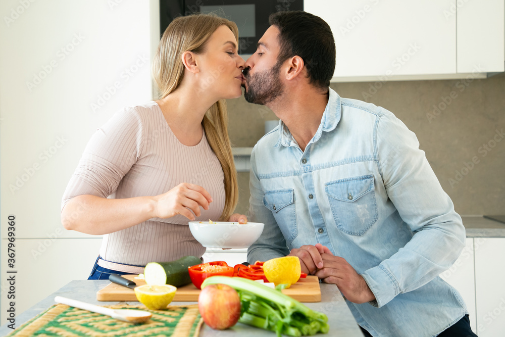 Happy sweet young couple kissing while cooking dinner together, cutting fresh vegetables on chopping board in kitchen, smiling and talking. Love and cooking concept