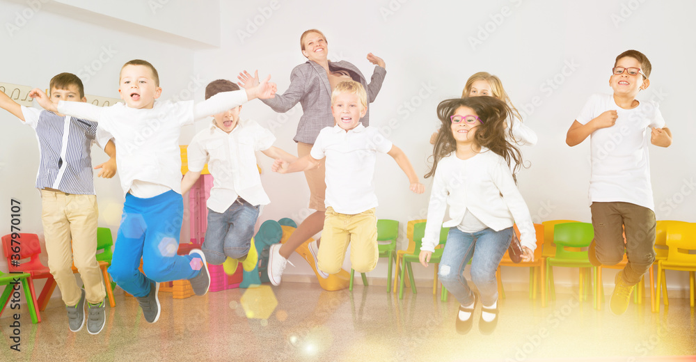 Obraz premium Group of happy cheerful children with their female teacher jumping together in a schoolroom