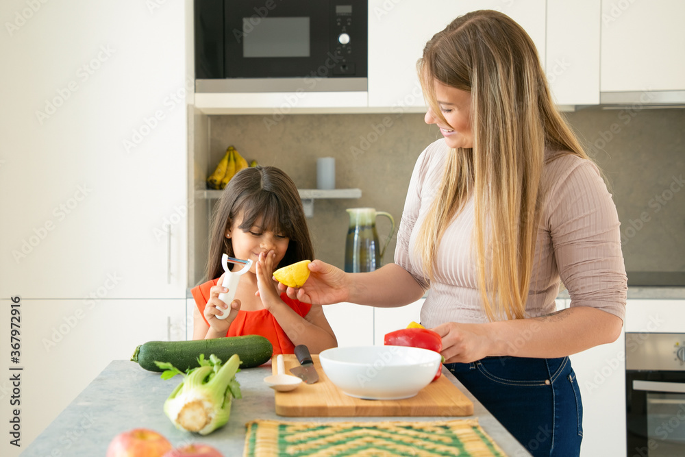 Happy mom and daughter having fun while cooking dinner together. Girl and her mother peeling and cutting vegetables for salad and lemon for dressing on kitchen counter. Family cooking concept