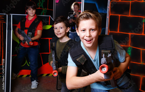 Emotional cheerful positive teen boy with laser pistol playing laser tag with friends on dark labyrinth