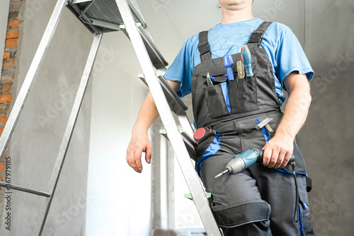 Guy in work clothes and with an electric screwdriver in his hand is standing on the stairs. Against the background of renovation in the house